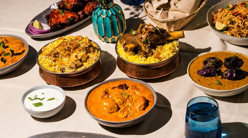 A table laden with various Indian dishes including biryani, curries, tandoori chicken, and noodles.